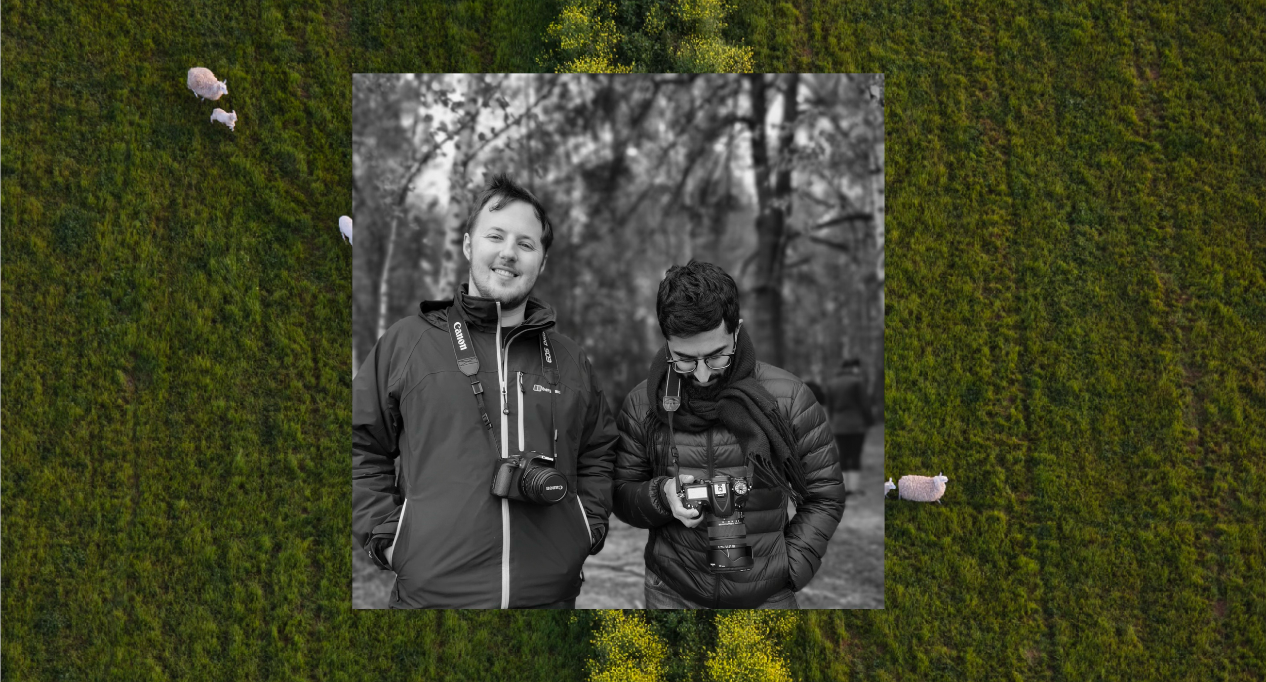 Two young men in jackets, one smiling at the camera with a camera hanging around his neck, and the other looking down at a camera he is holding, are standing outdoors in a park or forest area with trees in the background.