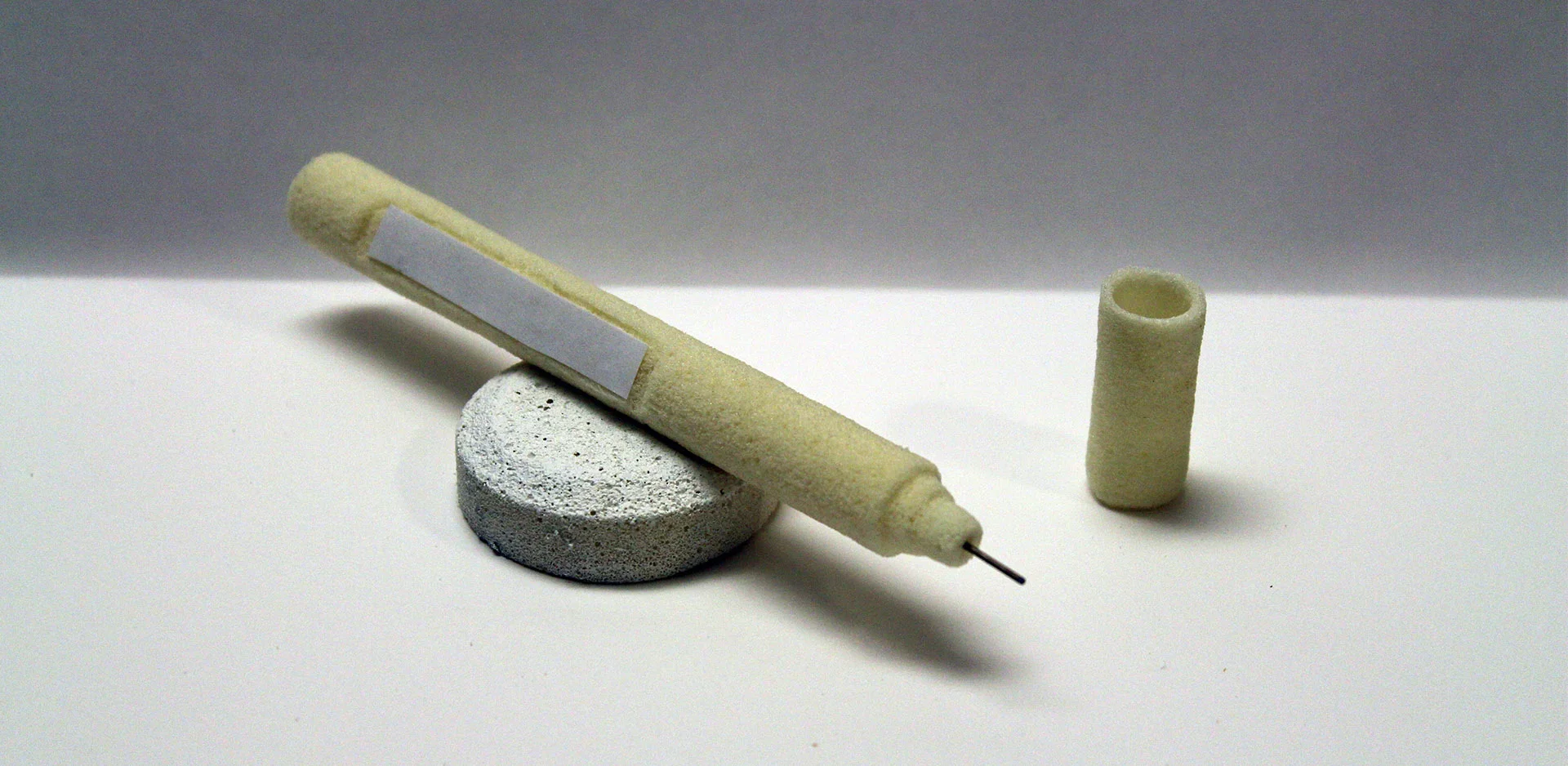 A plaster cast of a human finger and a small hollow tube, both white with a textured surface, are placed on a white surface with a gray background.