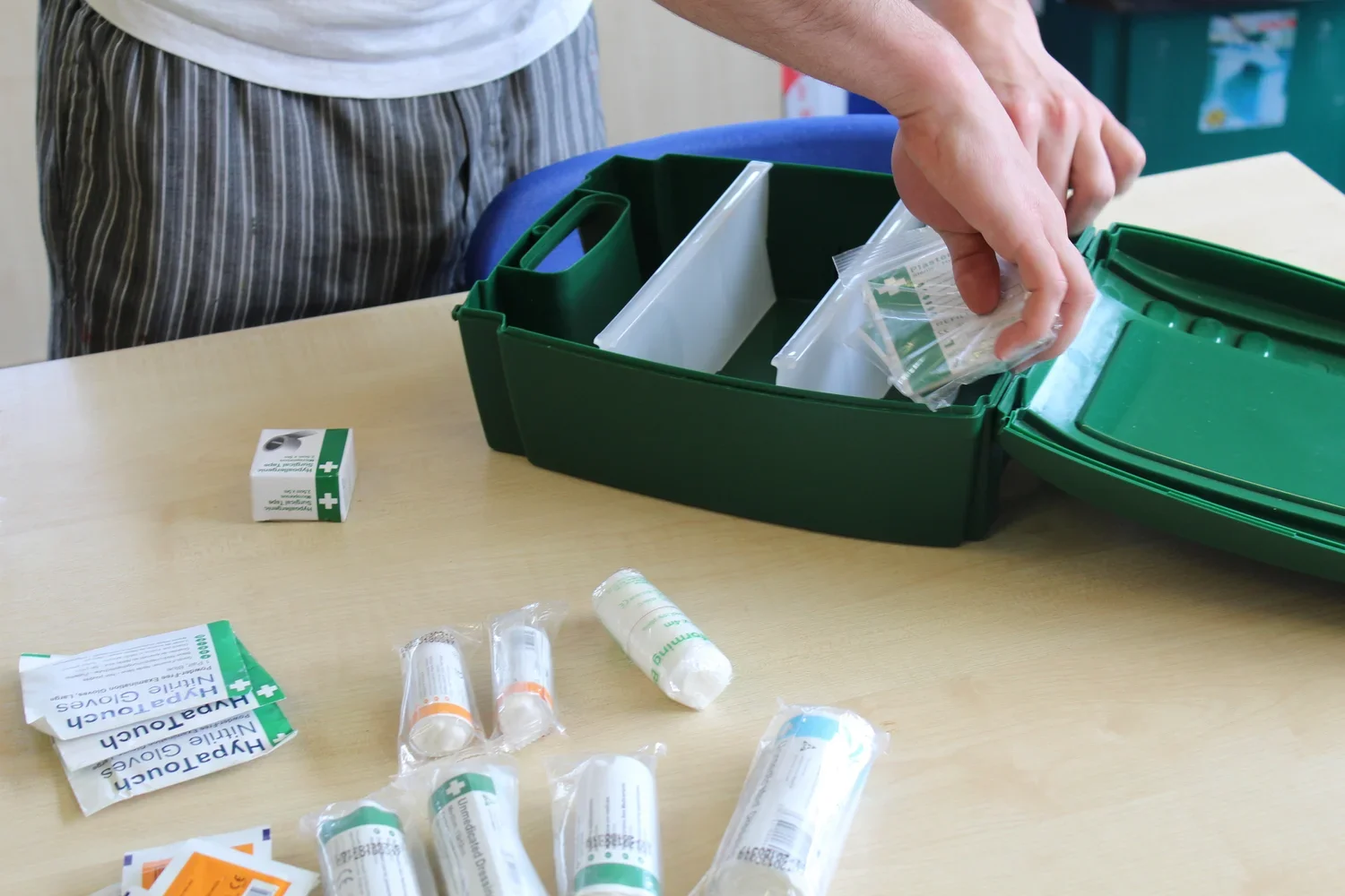 Person organizing a first aid kit with various medical supplies on a table, including bandages, ointments, and small pharmacy packets. The kit is green and the person's face is not visible.
