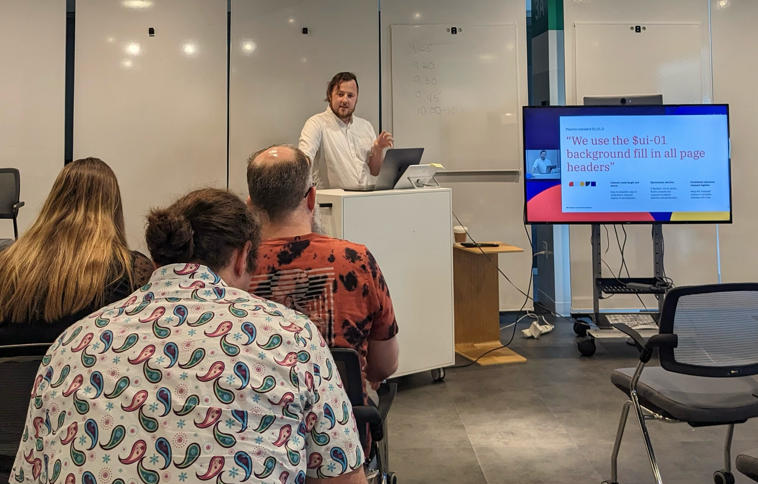 A man giving a presentation to an audience in a modern conference room. The presenter stands behind a white podium with a laptop, in front of a whiteboard. A large screen displays a slide with text about background fill and page headers. Four people are seated, listening to the presentation, with one wearing a colorful patterned shirt.