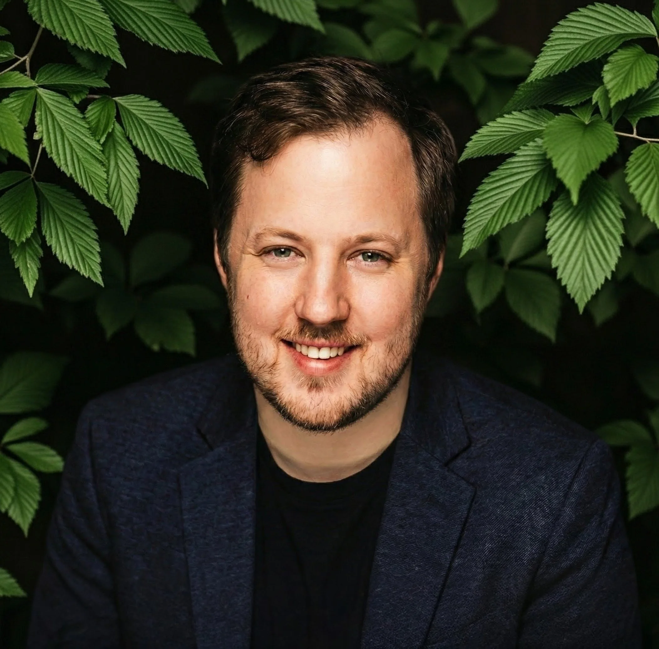 A man with light-colored hair and a beard smiling, sitting in front of green leafy plants, wearing a dark blazer and black shirt.