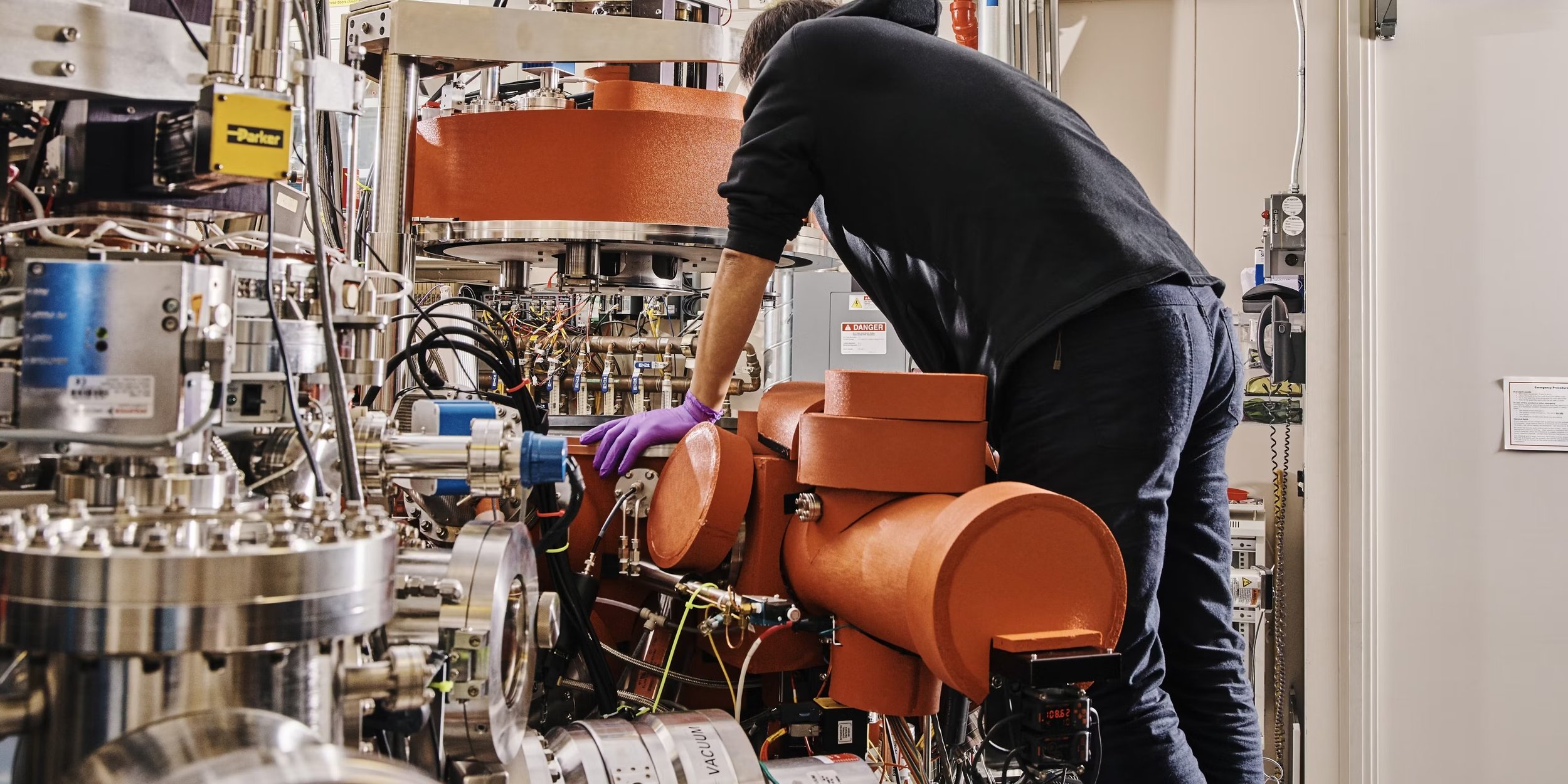 A scientist or engineer working with a large, complex scientific instrument or machinery in a laboratory, wearing purple gloves and bending over to examine or adjust the equipment.