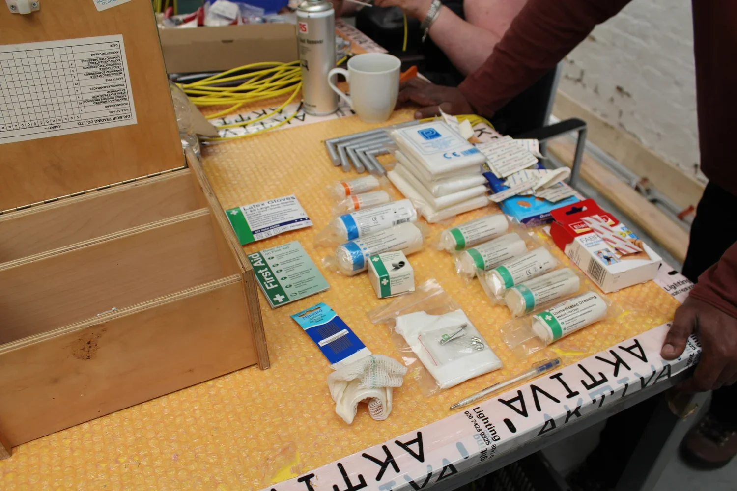 A table with various medical supplies including bandages, gloves, antiseptic wipes, a syringe, and a first aid kit, with a person reaching over.