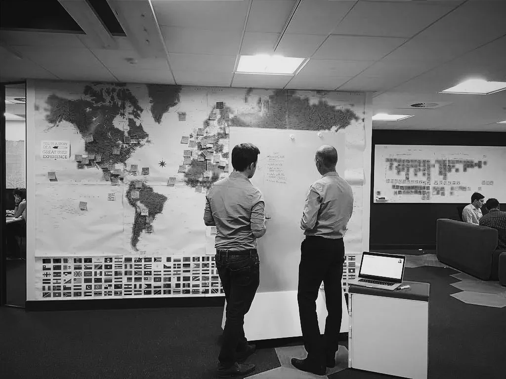 Two men in business casual clothing standing in front of a world map with notes and sticky notes, engaged in discussion in an office setting, with a laptop on a small table nearby and other people working in the background.