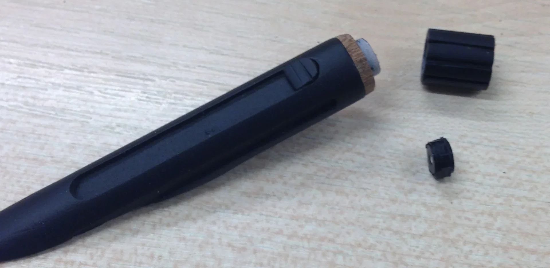 A black vape pen with a wooden top and three removable black parts lying on a light wooden surface.