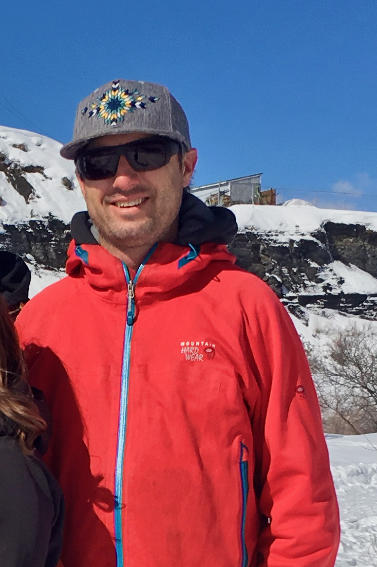 A man wearing sunglasses, a gray hat with a colorful star pattern, and a red Mountain Hard Wear jacket, standing outdoors in a snowy, mountainous area under a clear blue sky.