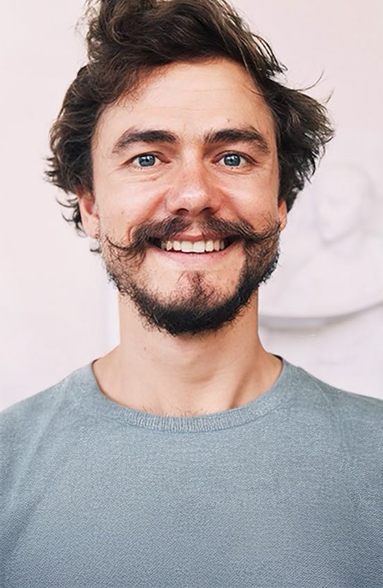 A smiling man with a beard and mustache, blue eyes, and tousled brown hair, wearing a light gray shirt.