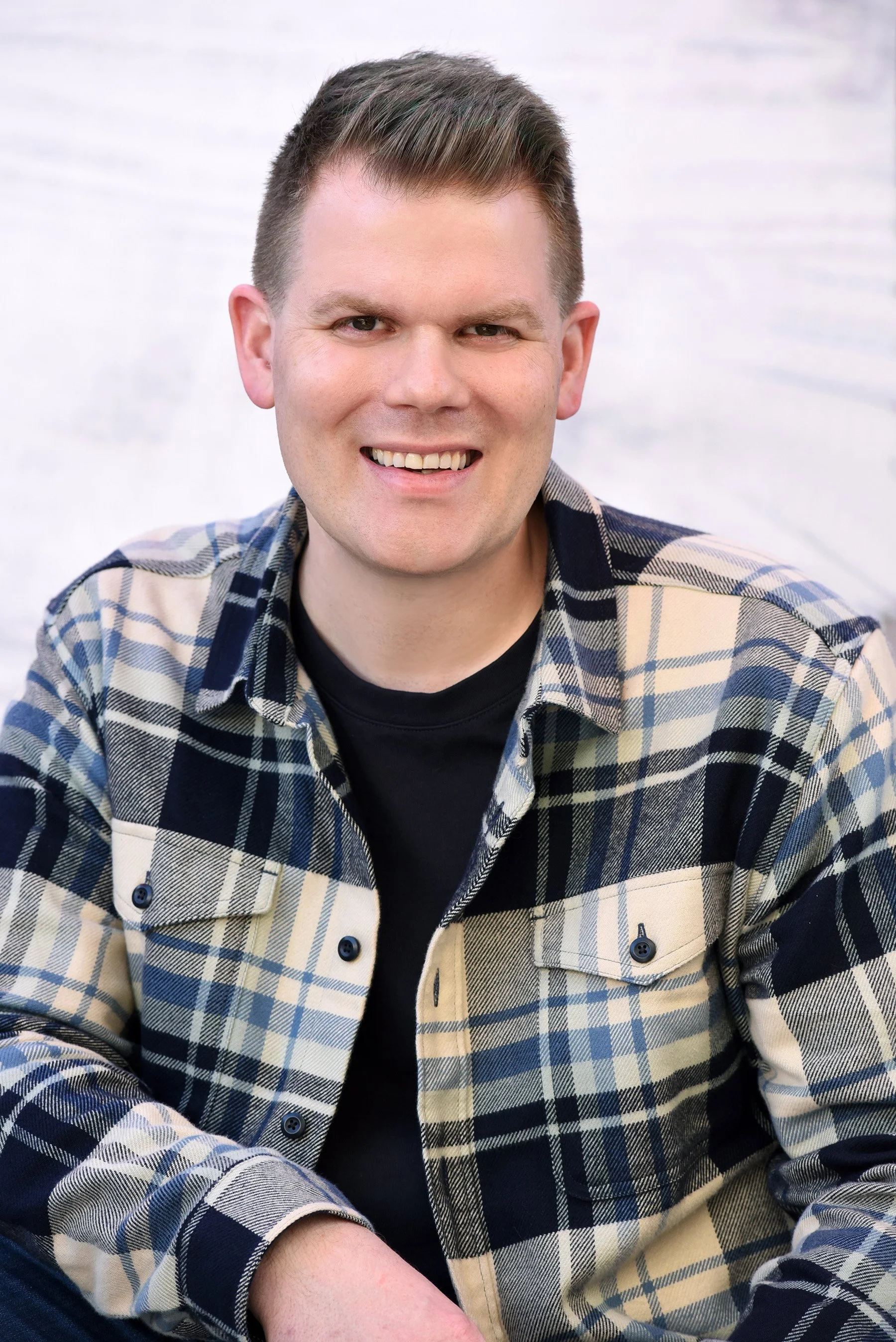 A smiling man wearing a plaid shirt and black t-shirt sitting against a white background.