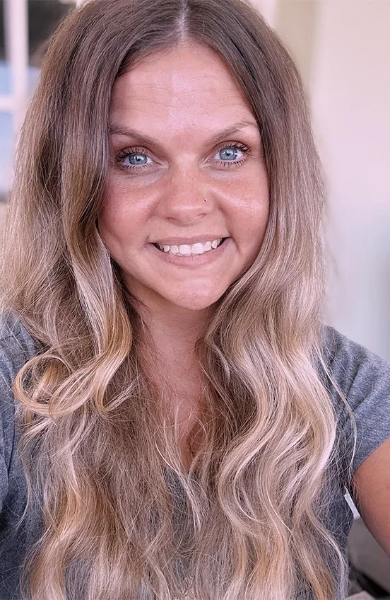 Close-up of a smiling woman with long, wavy blonde hair and blue eyes, wearing a gray shirt.