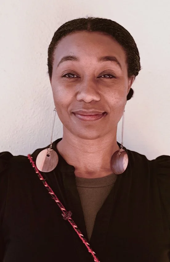 A woman with dark hair styled back, wearing large round earrings and a black top, standing against a plain wall, smiling subtly.