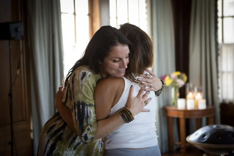 Two women hugging each other warmly indoors, with a background of curtains, candles, and a vase of flowers.