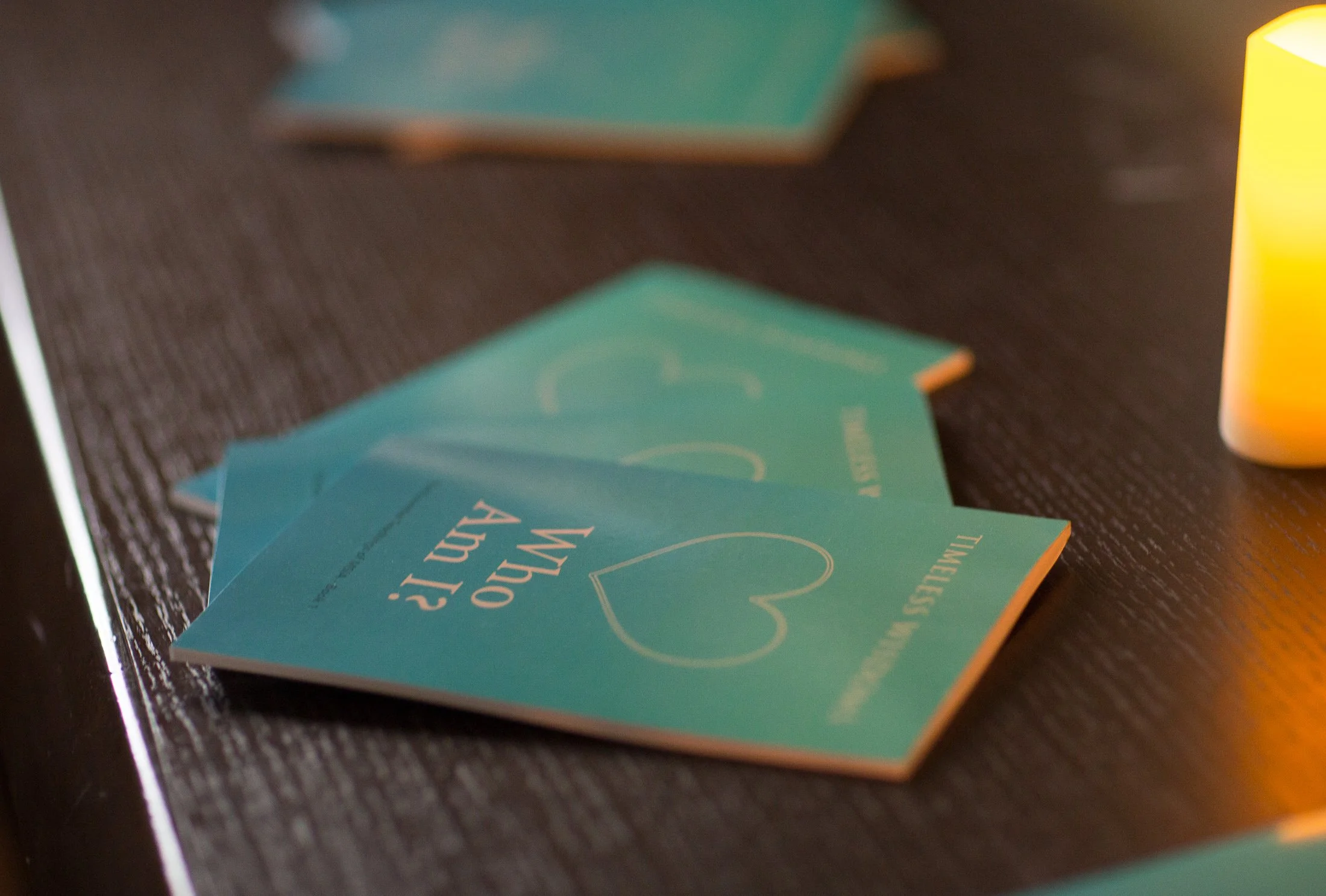 Three teal-colored cards with the words "Who am I?" and a heart outline, resting on a dark wooden surface, with a warm yellow light illuminating part of the scene.