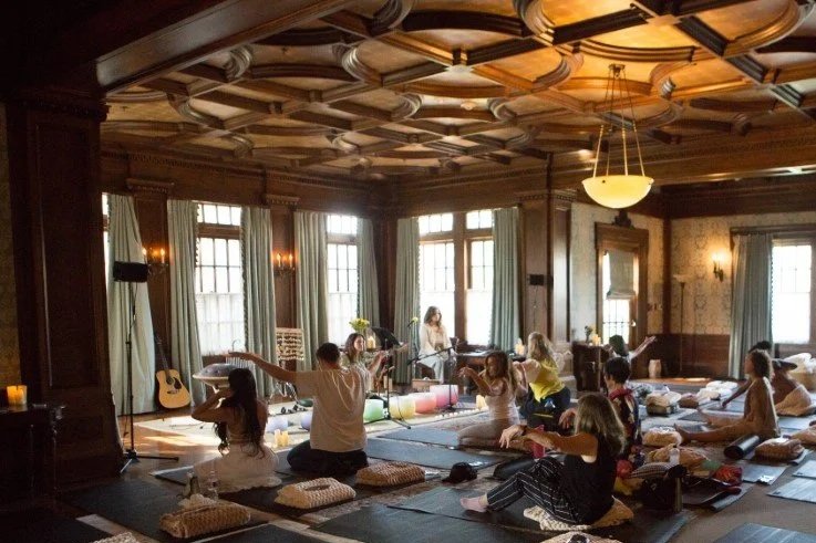 People participating in a yoga or meditation session in a large, well-lit room with wooden paneling, large windows, and a decorative ceiling. Participants are seated on cushions facing a woman standing at the front, possibly the instructor, with various yoga props and instruments around.