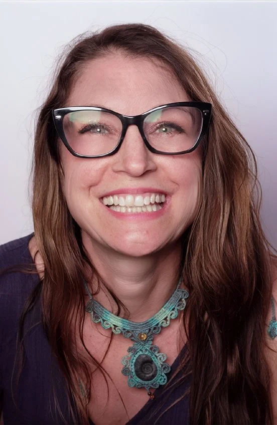 A woman with long brown hair, wearing black-rimmed glasses and a blue necklace, smiling broadly.