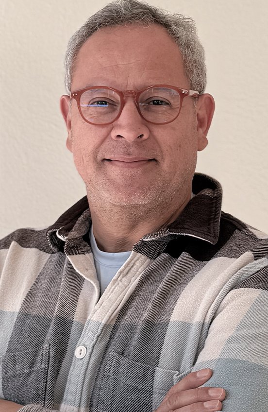 A smiling man with glasses wearing a plaid shirt standing in front of a plain wall.