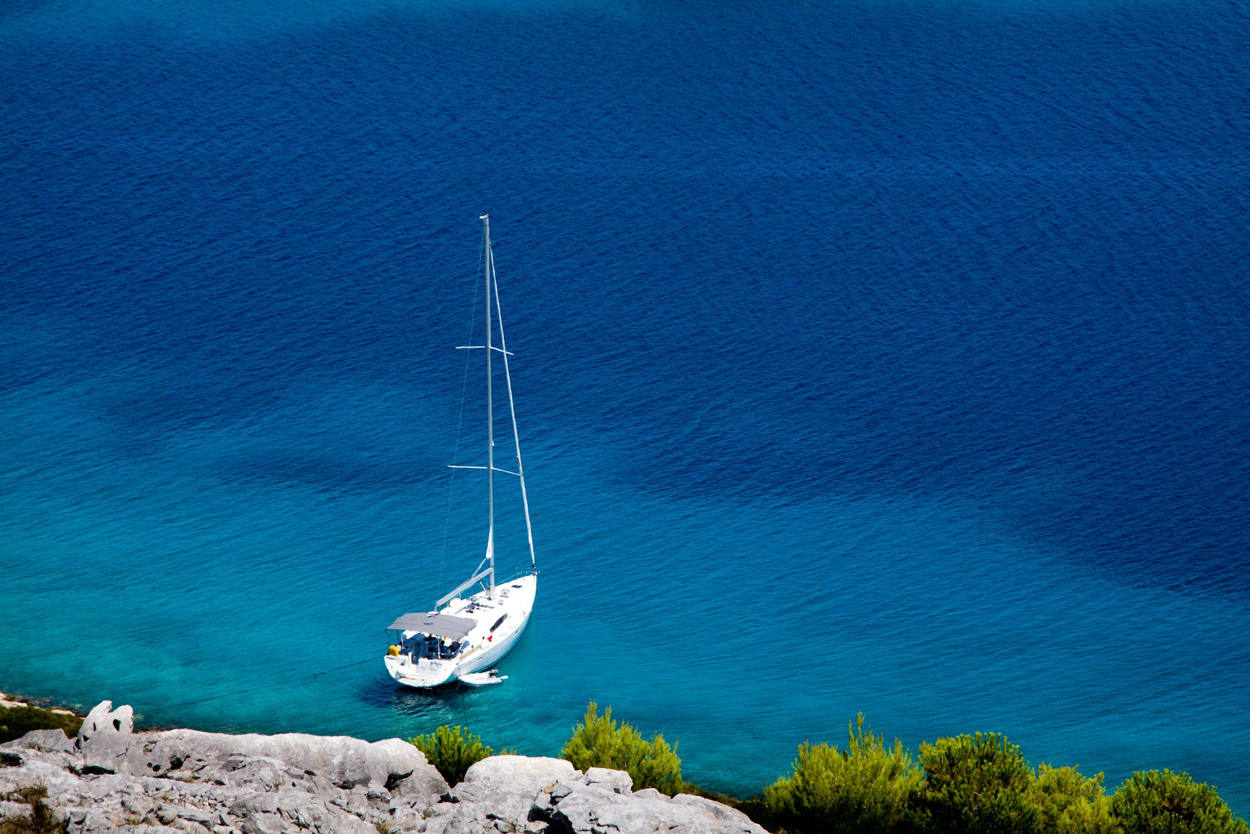 A white sailboat navigating clear turquoise waters near a rocky shoreline with green vegetation.