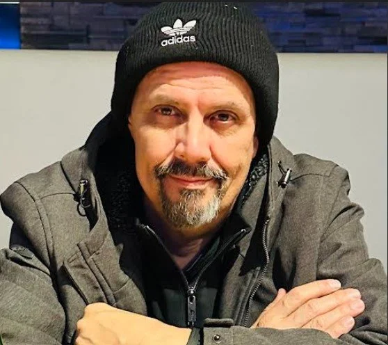 Mark Mazotti wearing a black Adidas beanie and gray jacket, sitting with arms crossed, indoors.