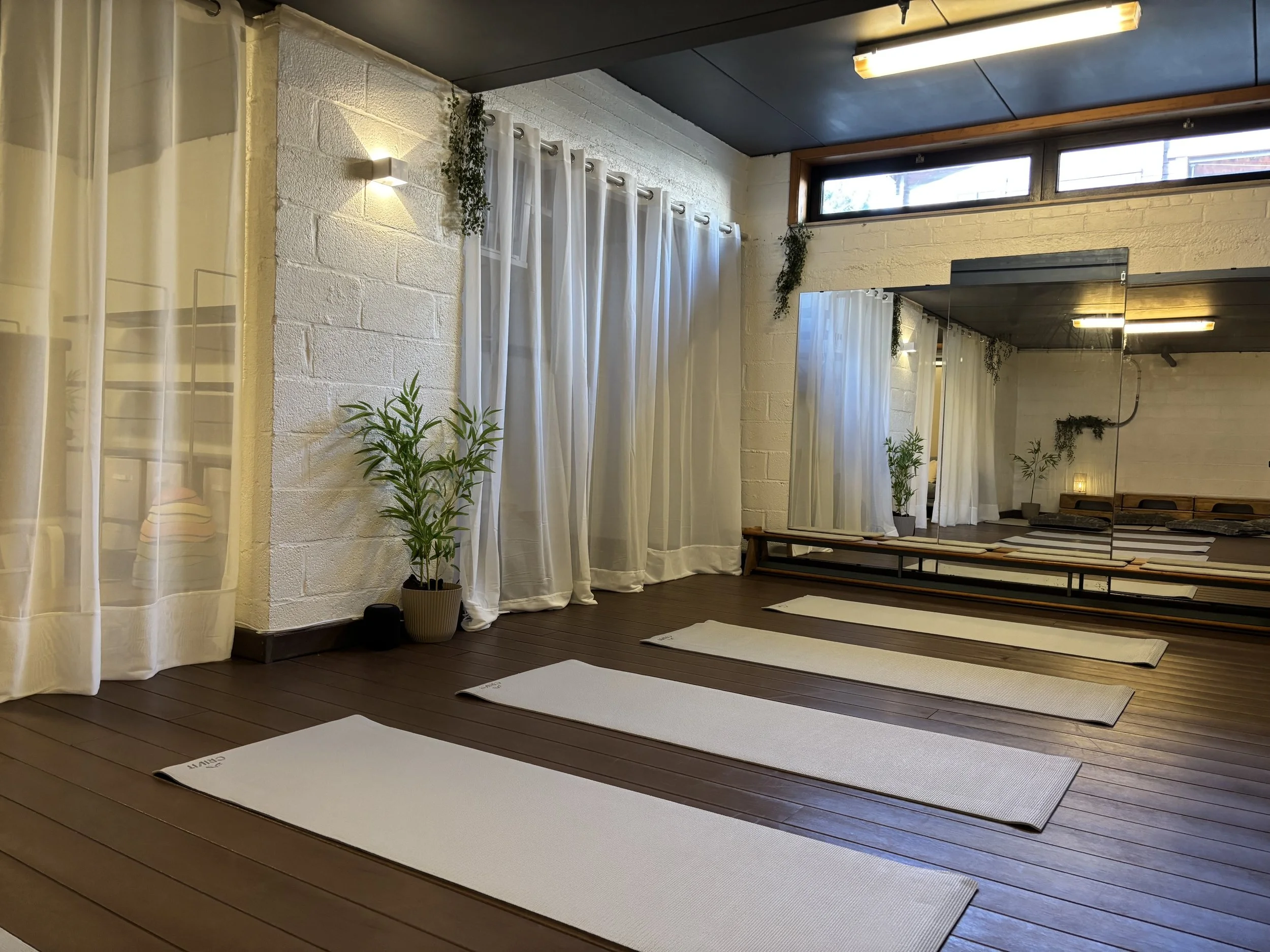 Binnenruimte van de_basis aangekleed voor een relaxatiesessie met witte gordijnen, planten, spiegels en yogamatten op de vloer.