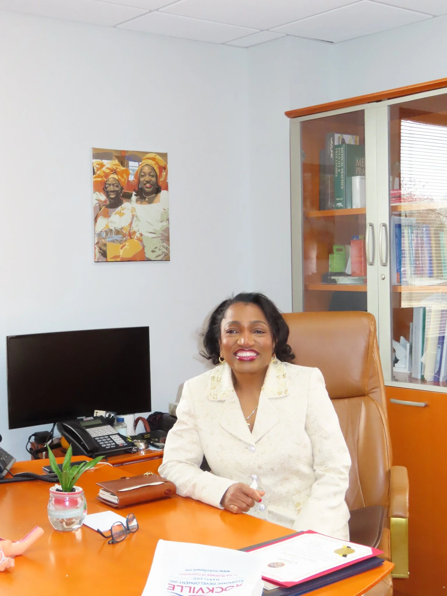 An African American woman smiling and sitting at an office desk with a computer, phone, and documents, in a professional setting.