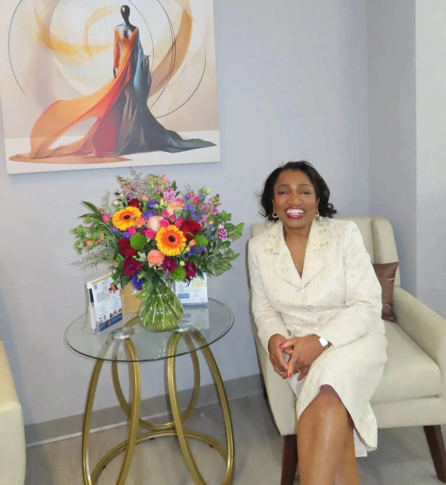 A woman dressed in a cream-colored suit sitting on a cream-colored sofa with a brown pillow, smiling at the camera. In front of her is a glass table with a large bouquet of colorful flowers in a glass vase, and some promotional materials. Behind her, there's a painting of a mannequin wearing a long, flowing gown, and the wall is painted light grey.