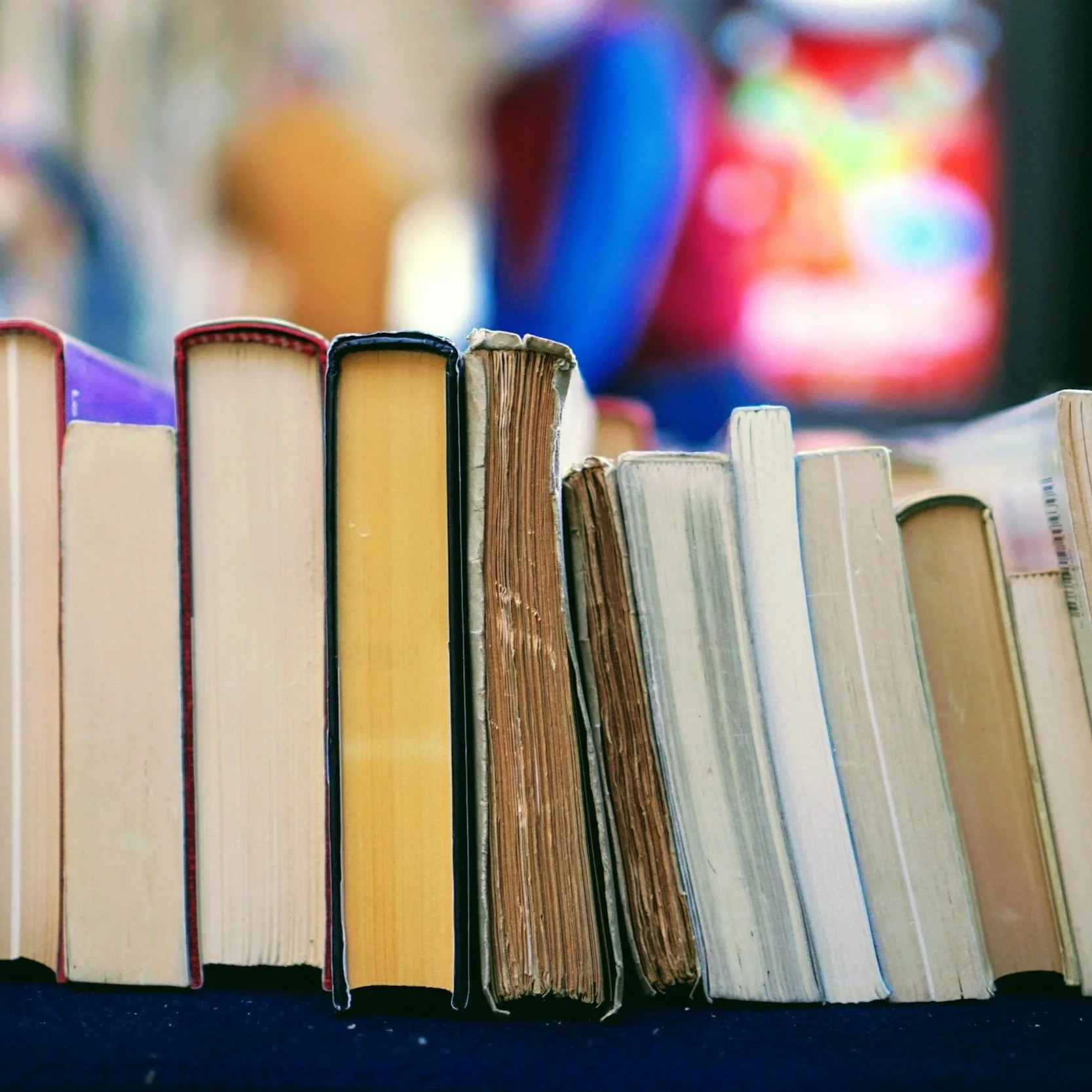 A row of used and new books standing upright on a shelf with a blurred colorful background.