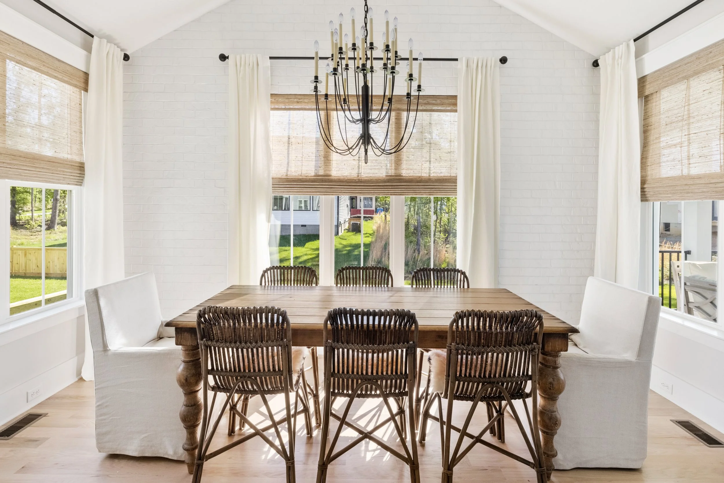 Dining room with large window, white brick walls, white curtains, wooden table, eight brown wicker chairs, and a black chandelier.