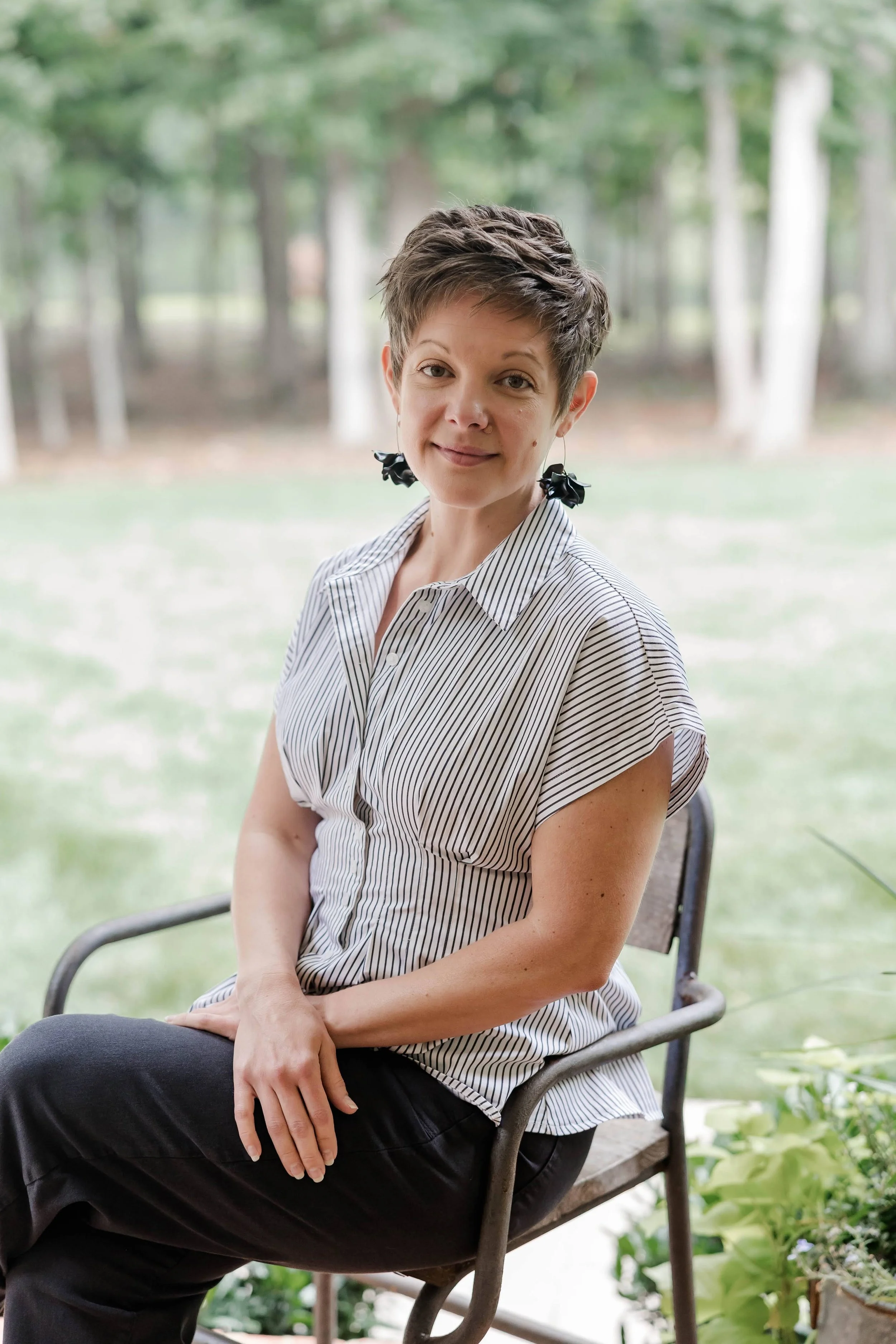 A woman with short brown hair, wearing a striped shirt and black pants, sitting on a gray chair outdoors near a pond with lily pads and green trees in the background.