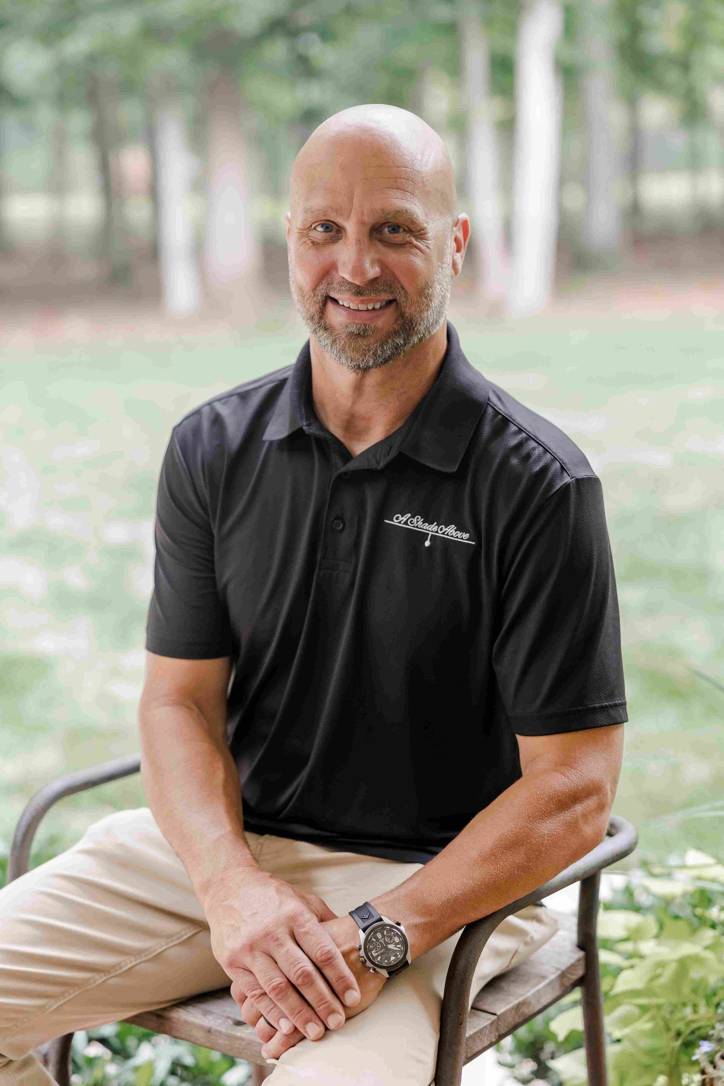 A smiling middle-aged man with a bald head and beard sitting on a wooden bench outdoors surrounded by greenery, wearing a black polo shirt and beige pants.