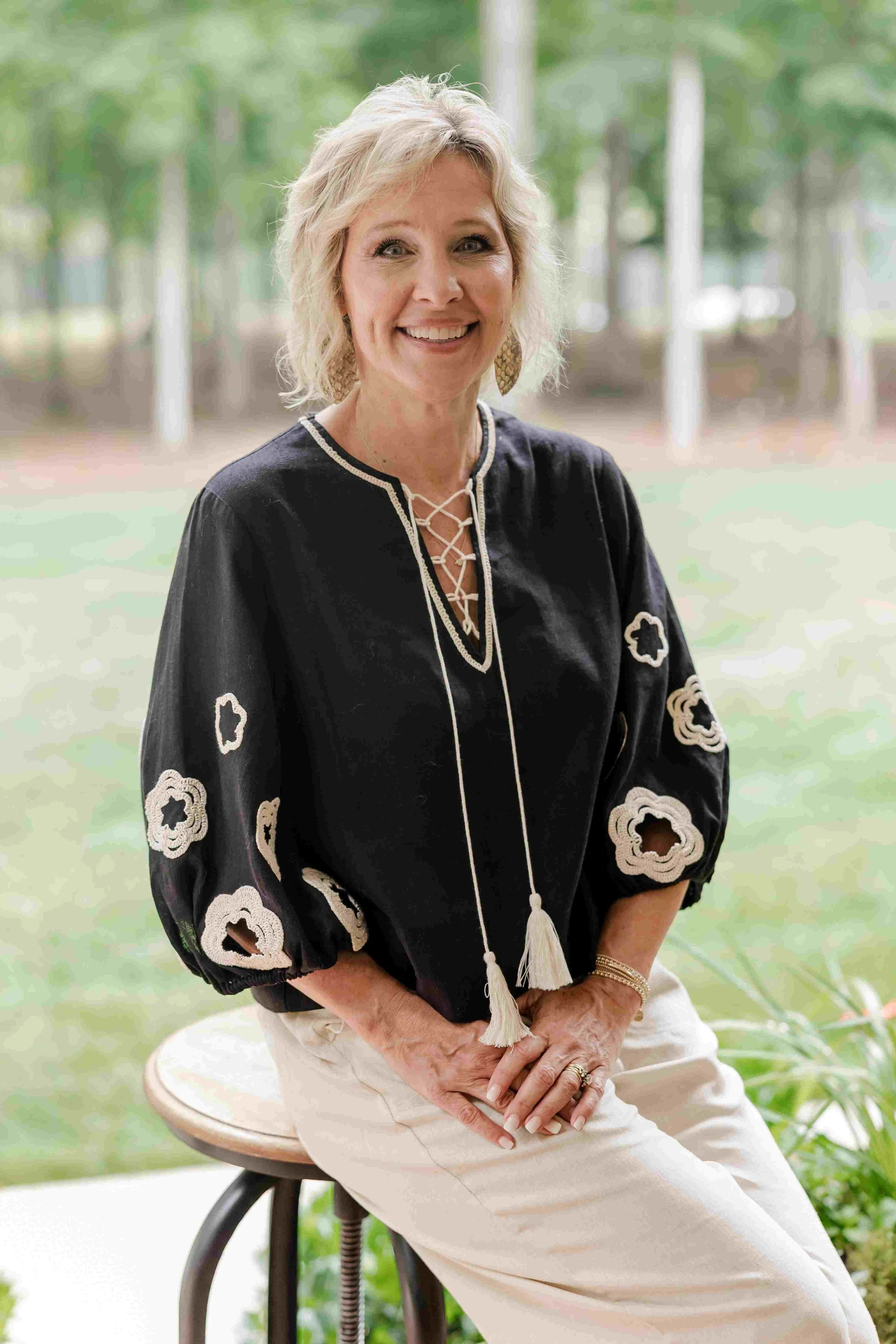 A smiling woman sitting on a stool outdoors with a blurred green background, wearing a black embroidered top and white pants.