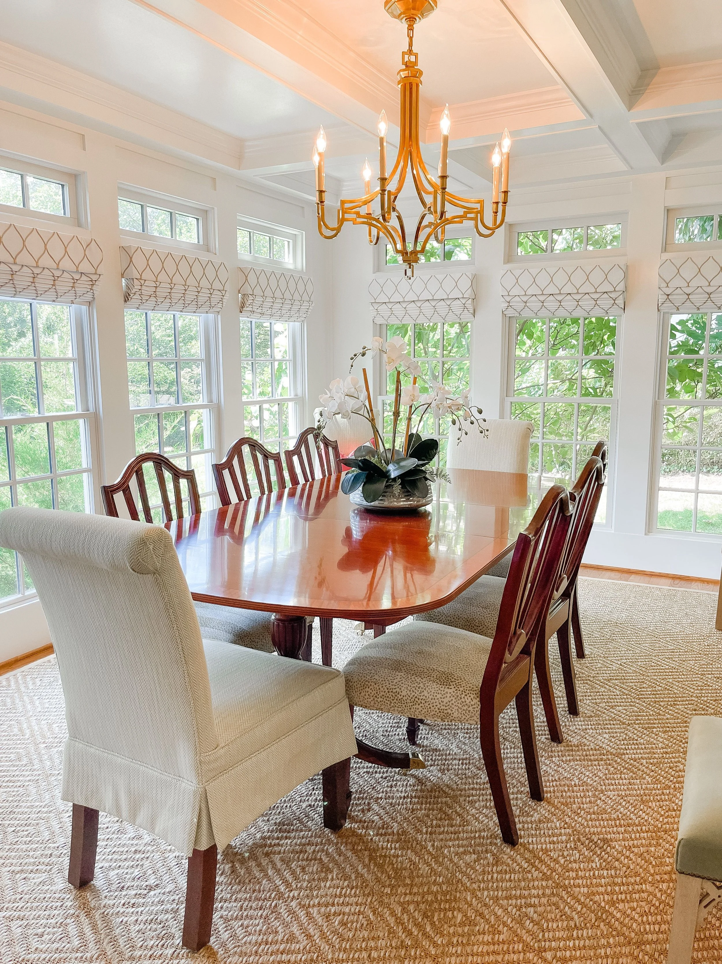 A dining room with a large wooden table, surround by white and patterned chairs. A decorative flower arrangement is on the table. The room has multiple windows with white frames and Roman shades. A gold chandelier hangs from the ceiling.