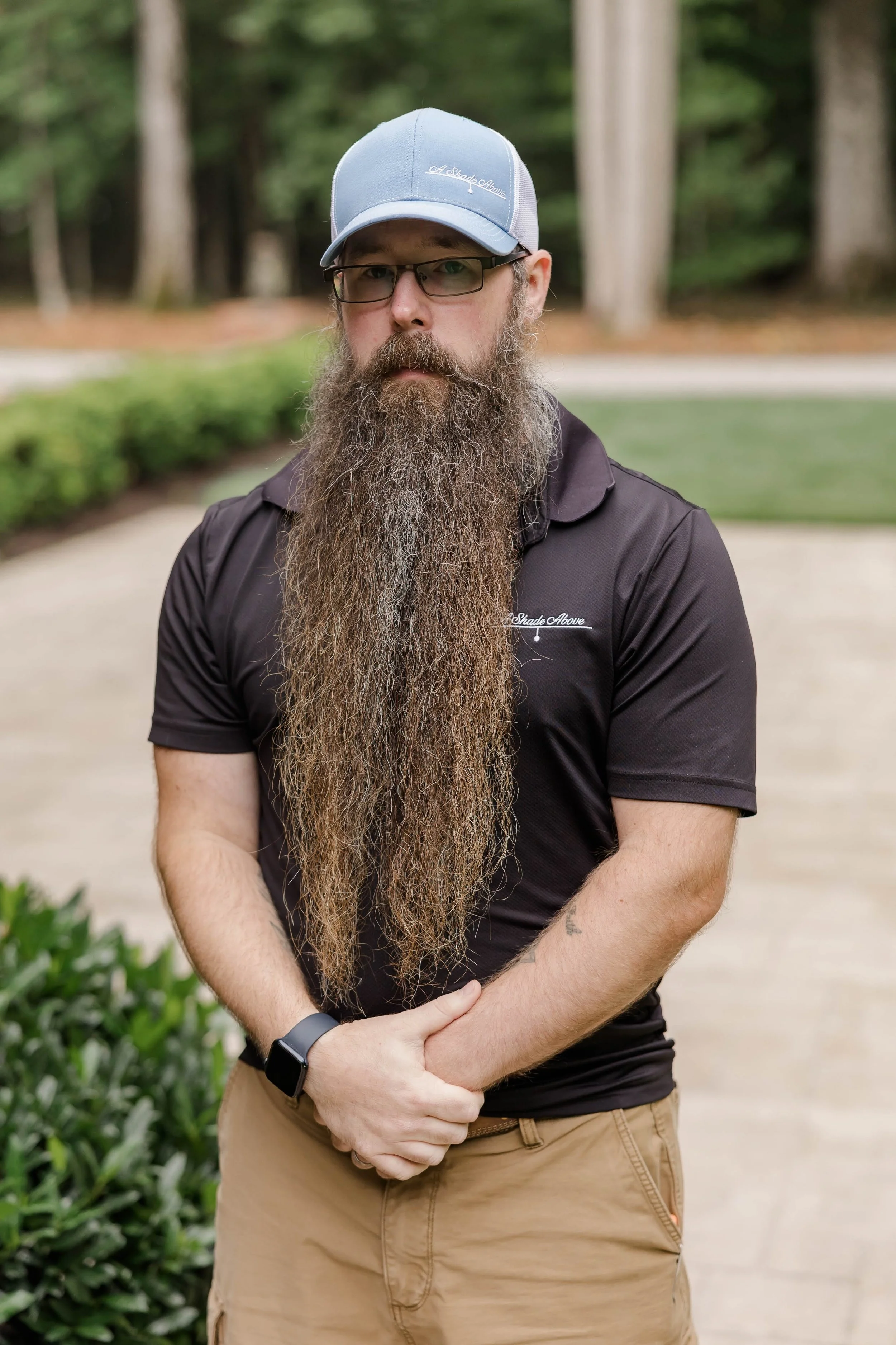 A man with glasses, a long beard, and a mustache standing outdoors in front of trees and bushes. He is wearing a light blue and white baseball cap and a black shirt with a logo on it. He has a black watch on his left wrist and is crossing his arms.