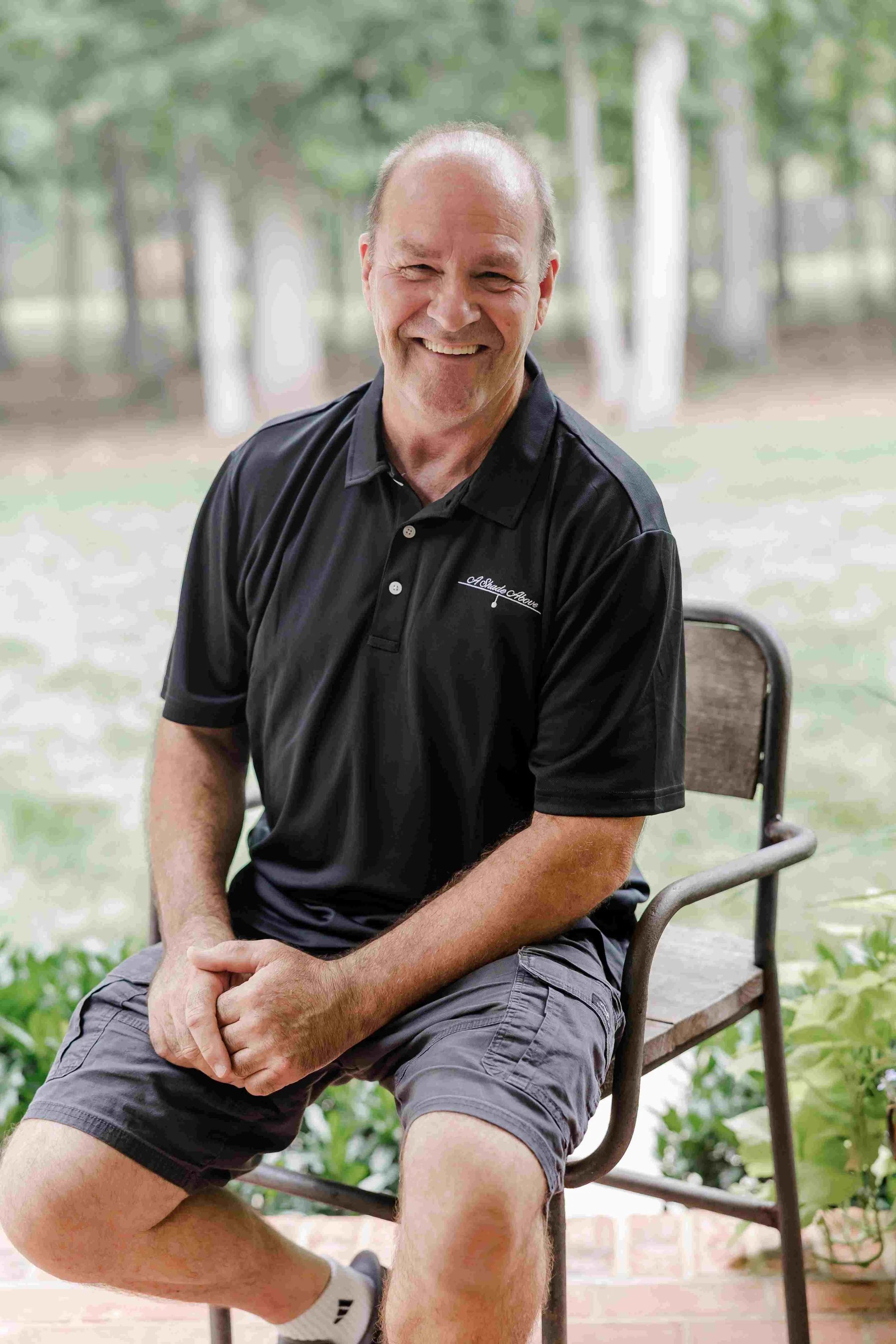 A smiling older man sitting outdoors on a park bench with trees and greenery in the background.