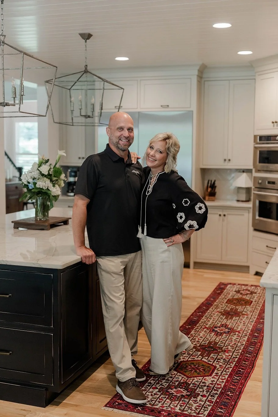 A man and a woman standing in a modern, white kitchen with white cabinets, stainless steel appliances, and a decorative runner on the wooden floor. They are smiling, with the woman having her arm around the man's shoulder and the man resting his hand on a marble kitchen island with a floral arrangement.