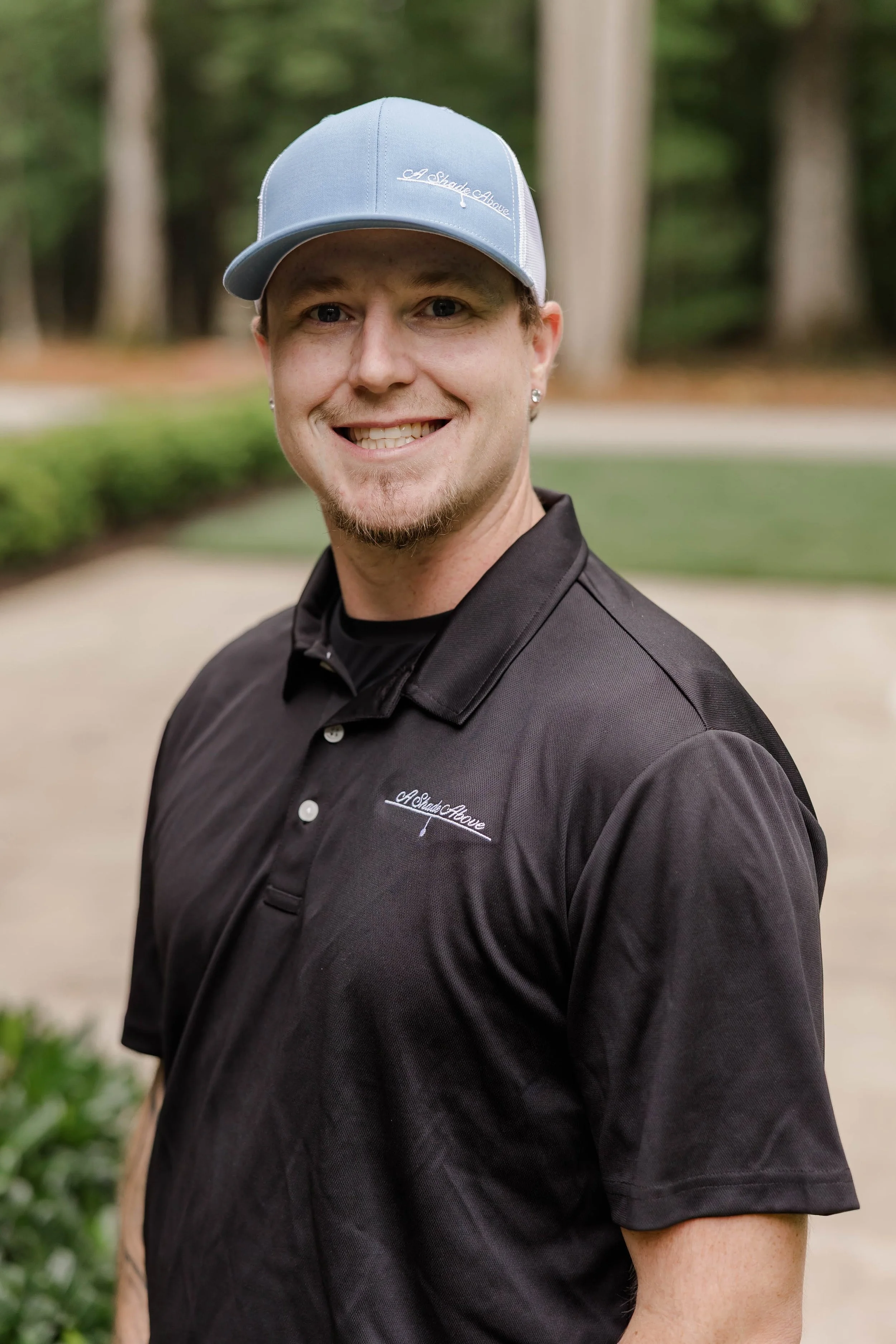 Man wearing a light blue cap and black polo shirt outdoors, smiling, with trees and greenery in the background.