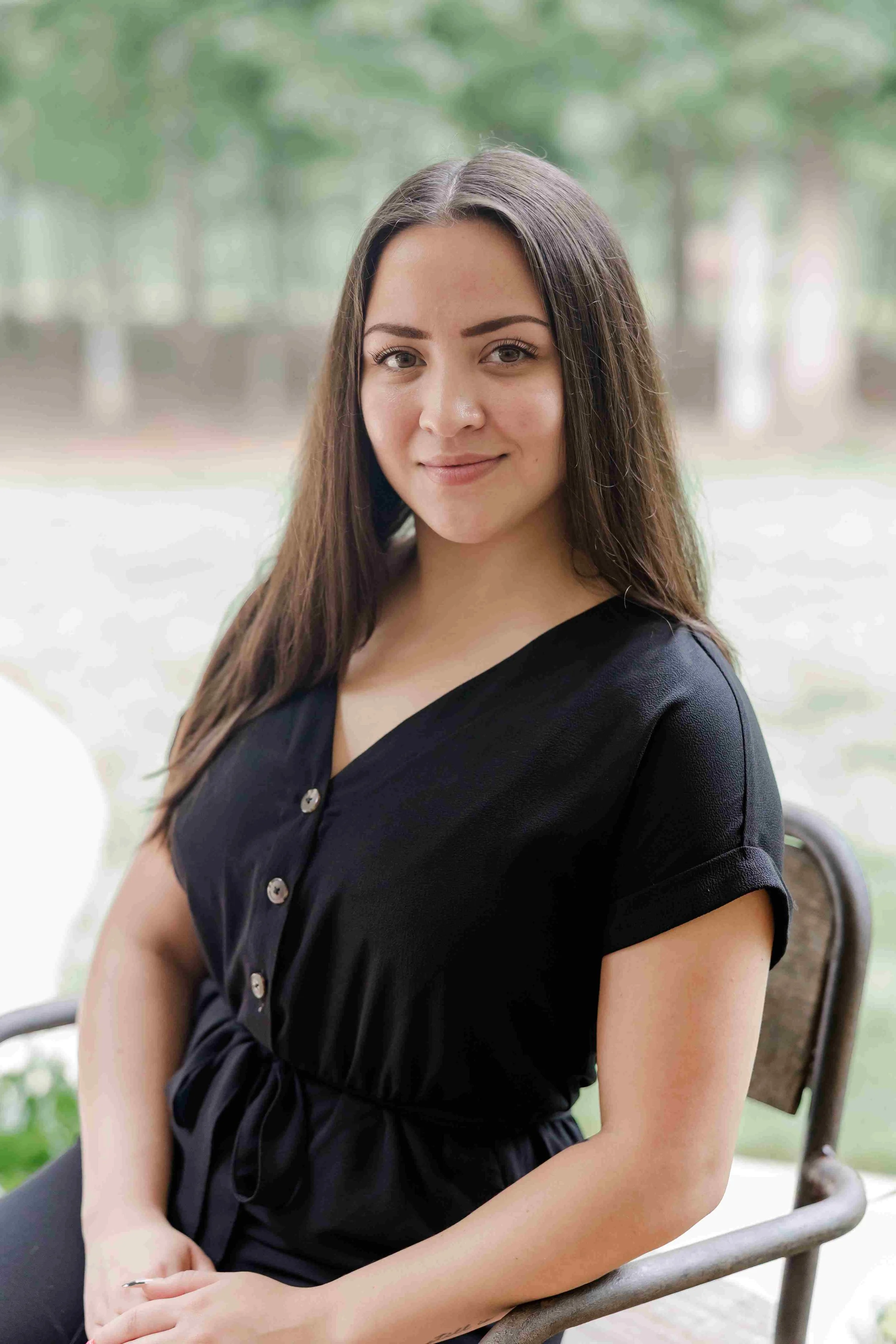 A young woman sitting outdoors on a metal bench, smiling at the camera with long brown hair, wearing a black button-up shirt.