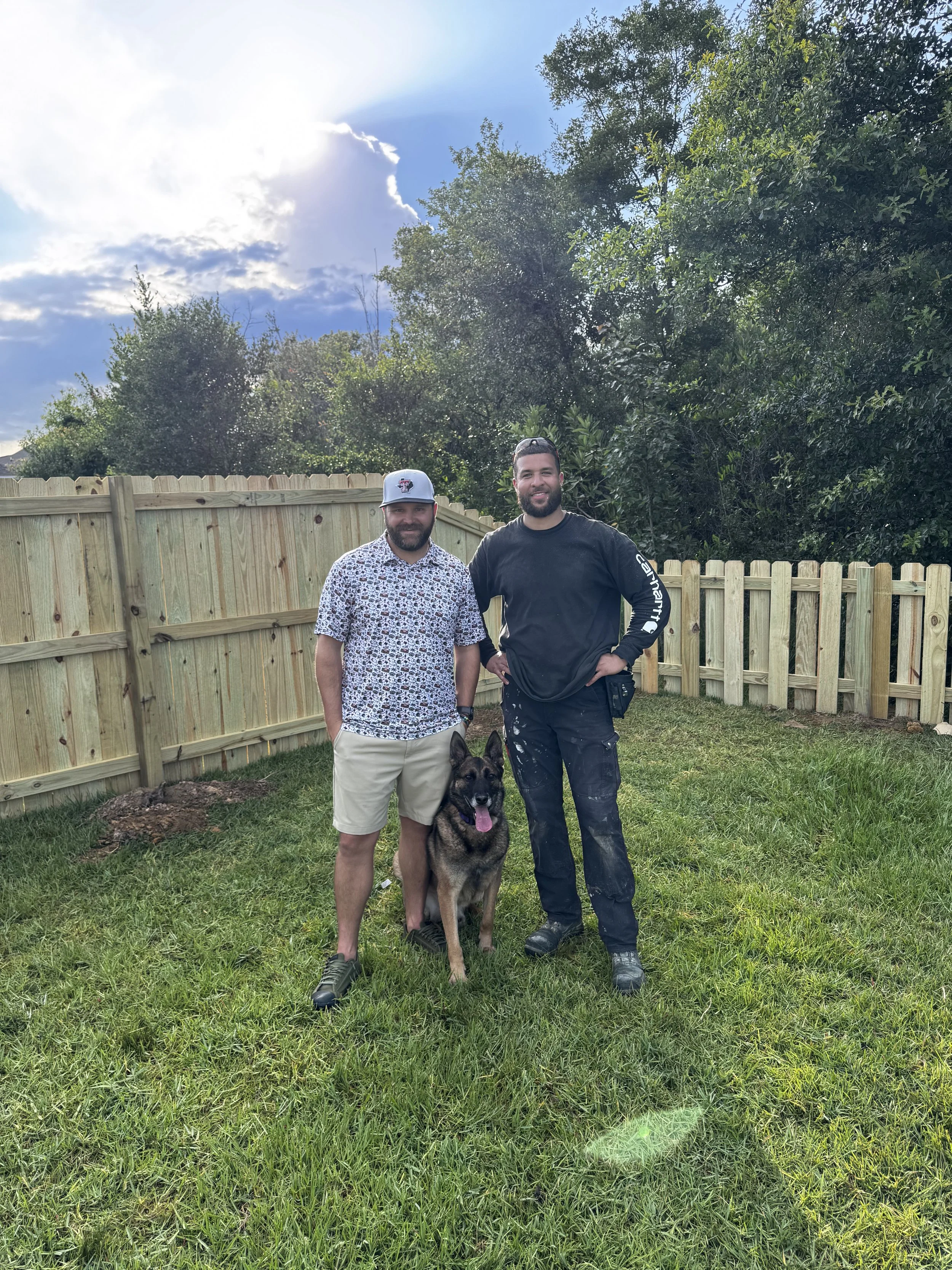 Two men and a German Shepherd dog standing on grass in a backyard with a wooden fence and trees in the background.