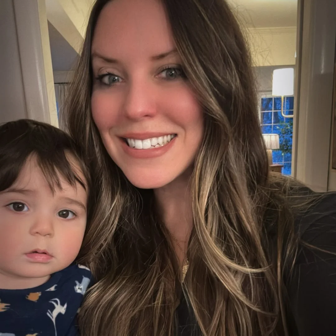 Woman with long, wavy brown hair smiling next to a young child with dark hair and a surprised expression, indoors in a warmly lit room.