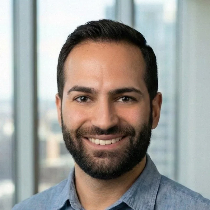 Portrait of a smiling man with dark hair and a beard, wearing a blue collared shirt, standing in front of large windows with a cityscape in the background.