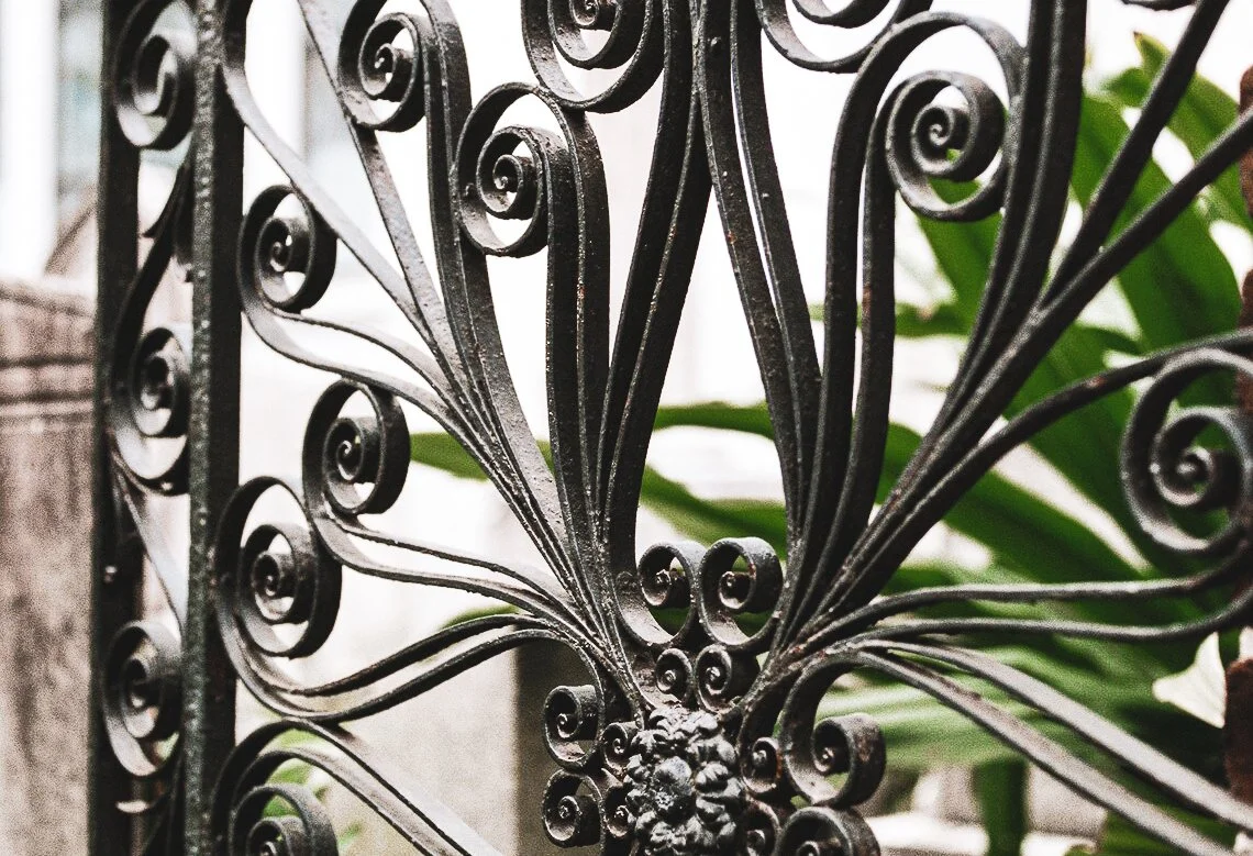 Close-up of an ornate black wrought iron fence with intricate scrollwork, with green leaves in the background.