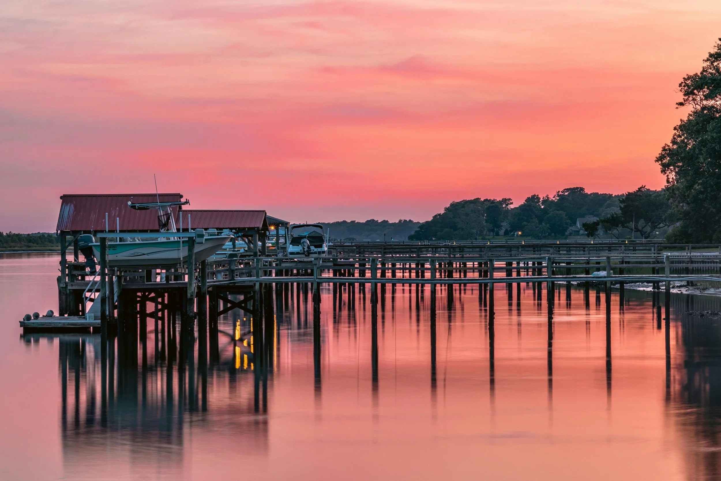 A peaceful scene of boats docked at a pier during sunset with pink and orange sky reflections on calm water.