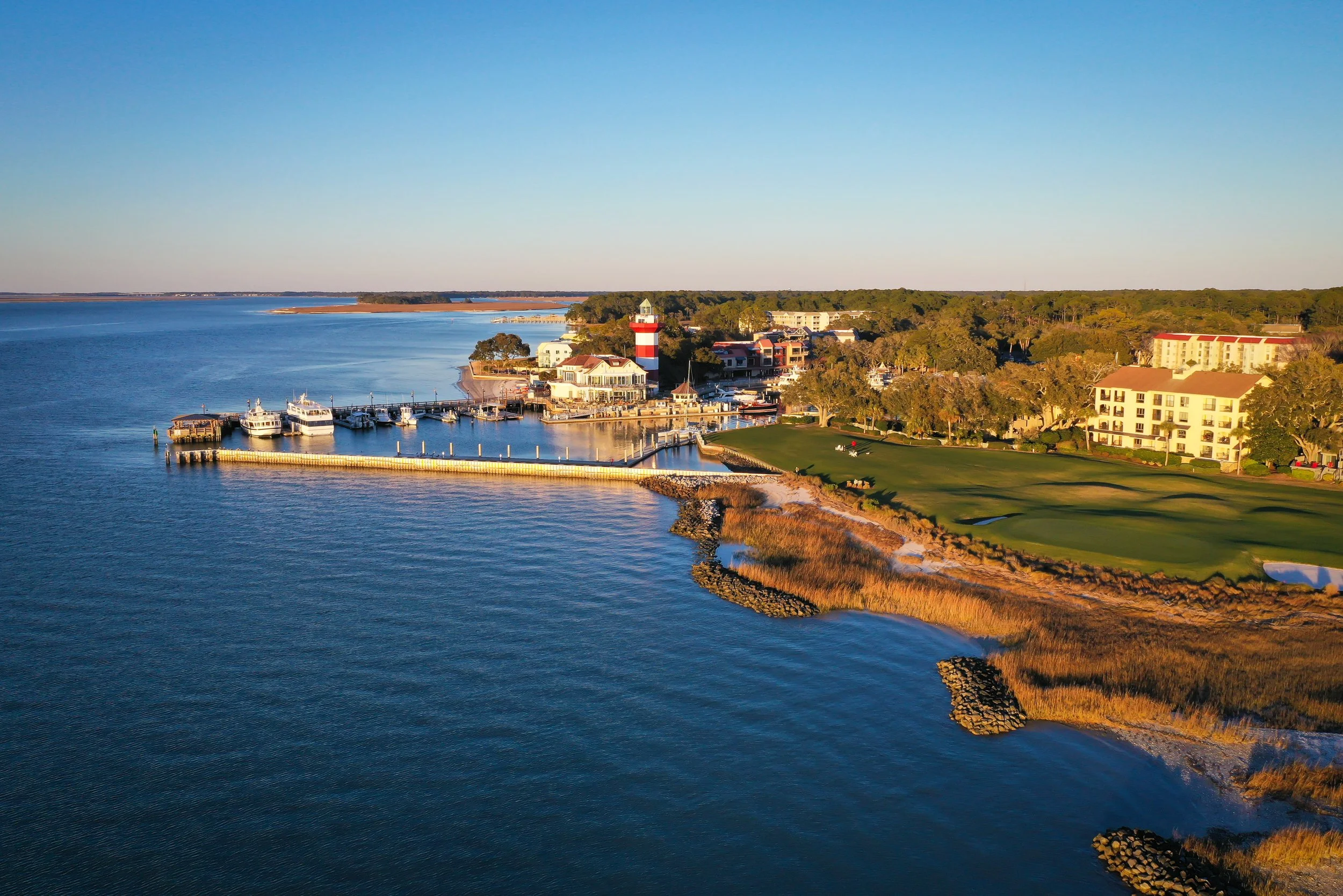 A waterfront scene with a marina, boats, a lighthouse, greenery, and buildings under a clear blue sky during sunset.