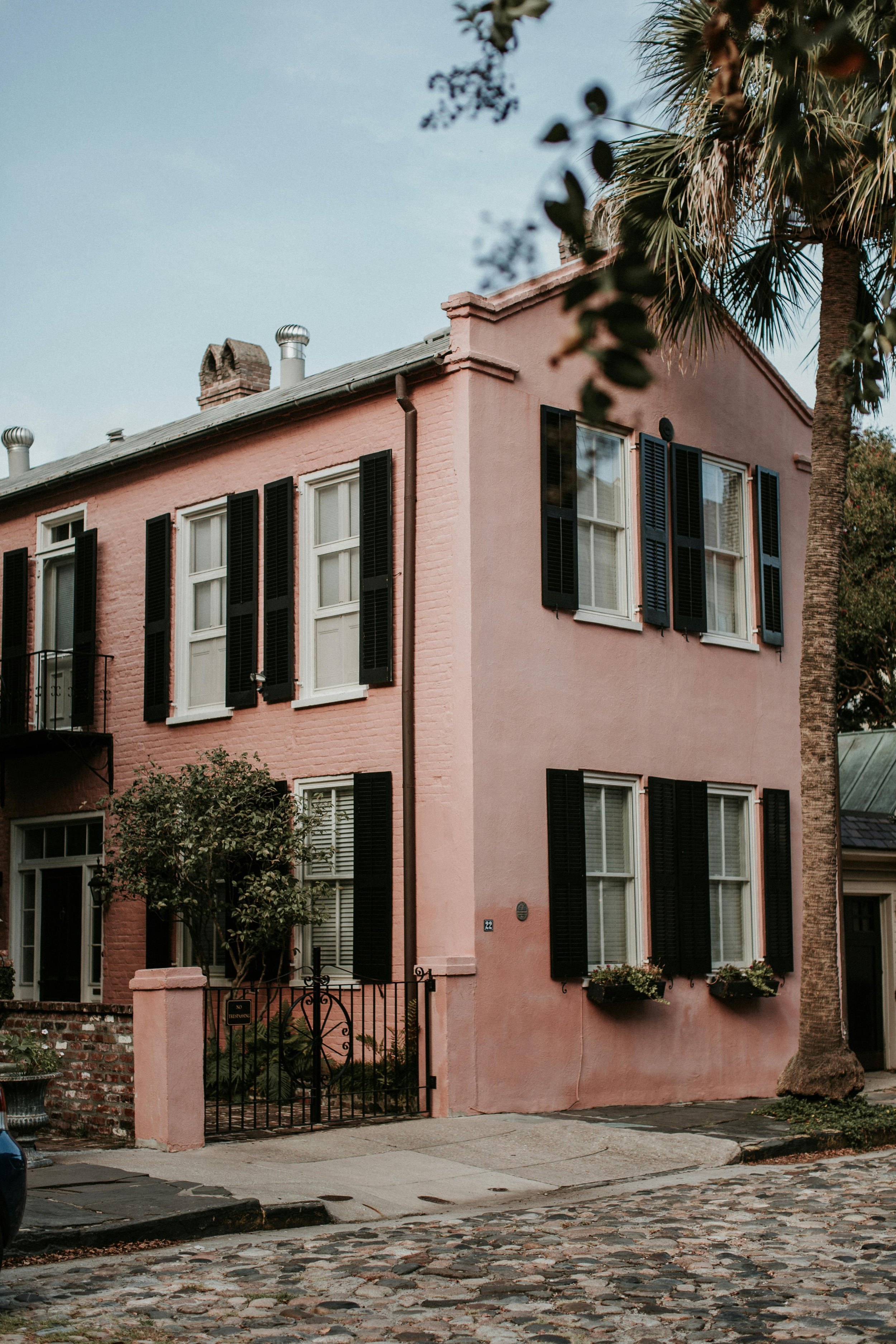 A pink three-story house with black shutters and large windows, a black iron gate, and some greenery. A tree is visible on the right side, and the house has a cobblestone alley in front.
