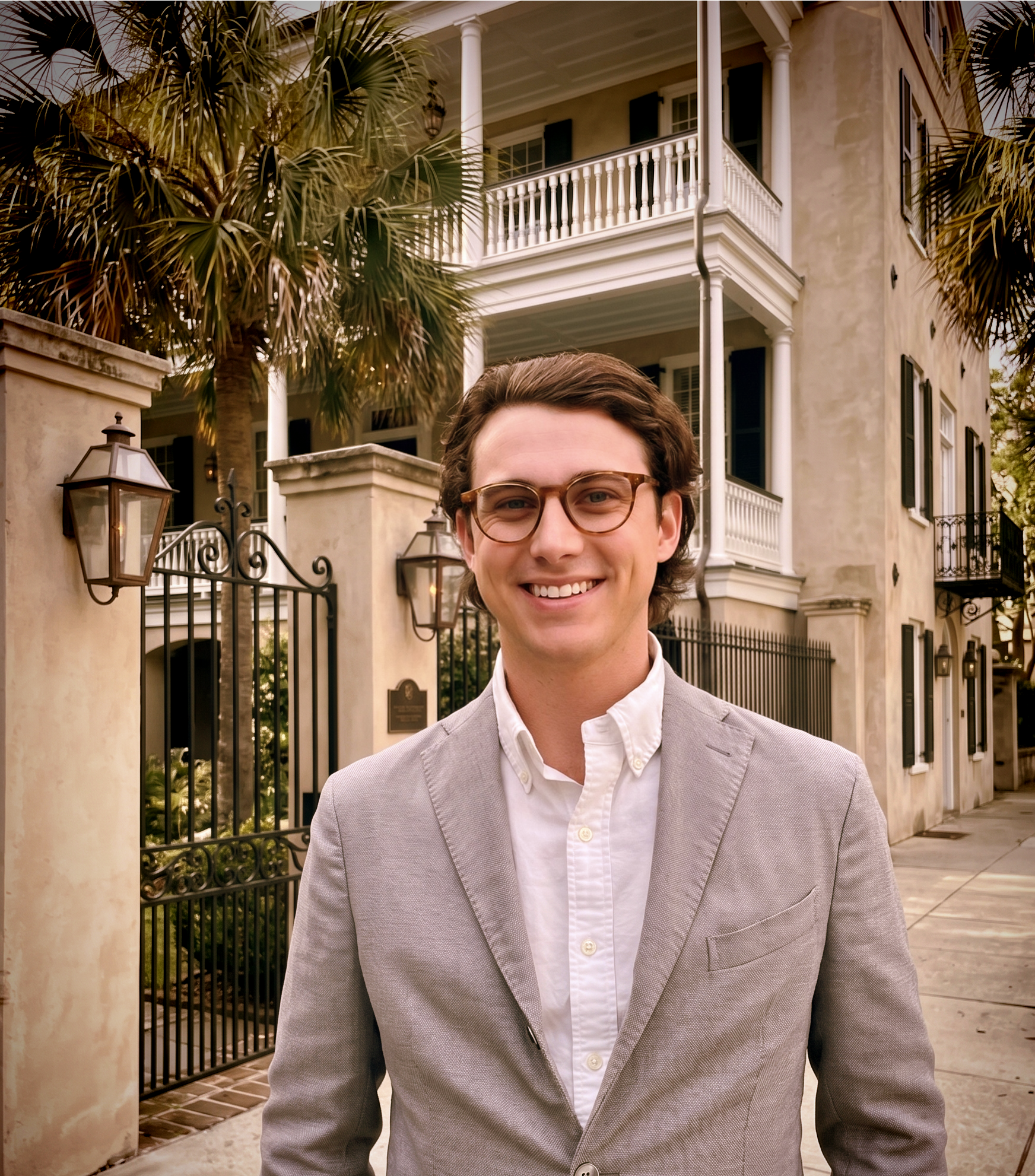 Half of a smiling man wearing glasses and a light gray suit, standing outside a large, cream-colored house with balconies and black shutters, surrounded by palm trees and outdoor lanterns.