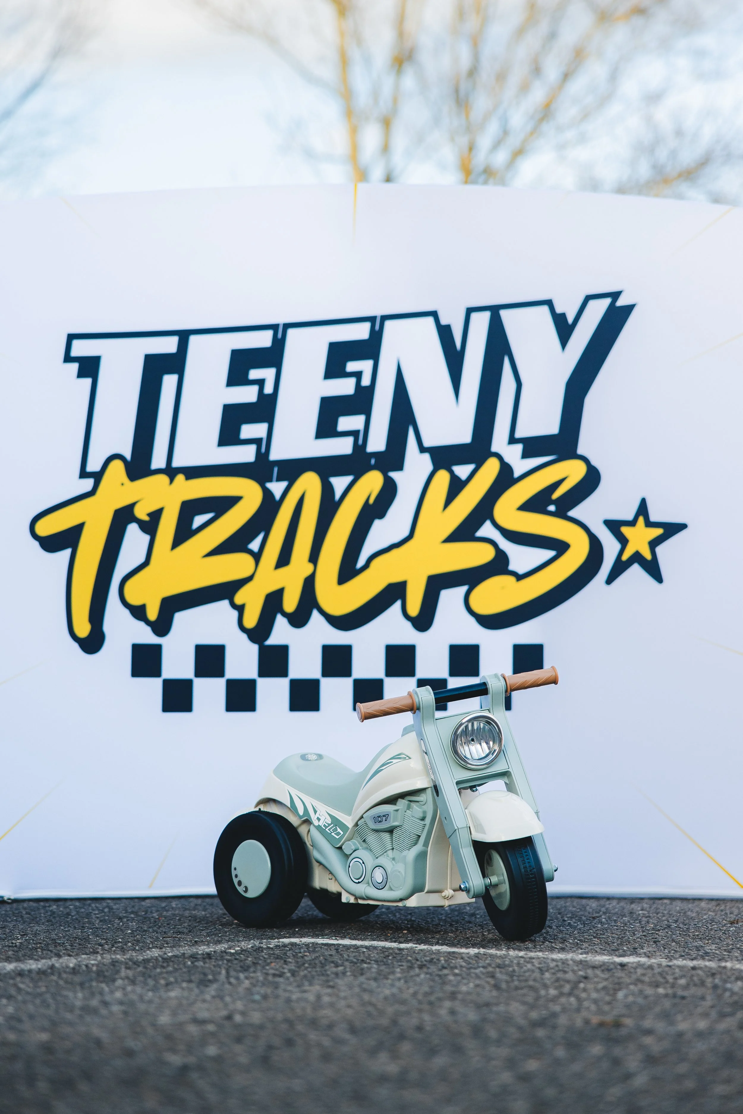 Small beige toy bubble motorcycle with black wheels. The scene is outdoors on asphalt with a faint reflection of the toy on the ground.