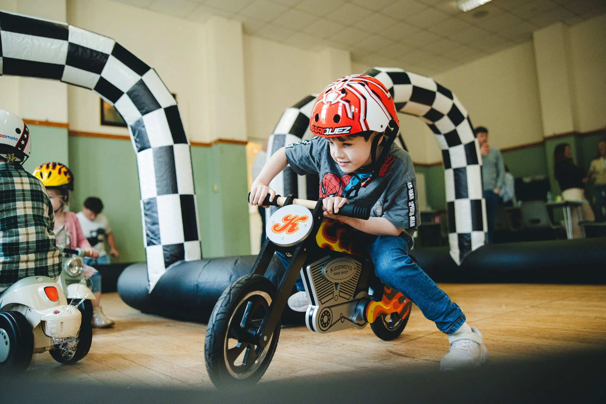 Young boy wearing a red helmet riding a black and orange toy motorcycle at an indoor play area with checkered archway and other children in the background.
