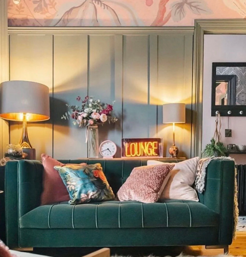 A cozy living room with a dark green velvet sofa adorned with multiple pillows, including a floral pillow, a pink pillow, and white pillows. Behind the sofa, there are two table lamps with beige shades, a vase of flowers, a round clock, and a neon sign that reads 'LOUNGE.' The room features muted green wainscoting on the walls, and there's a decorative floral or botanical painting above.