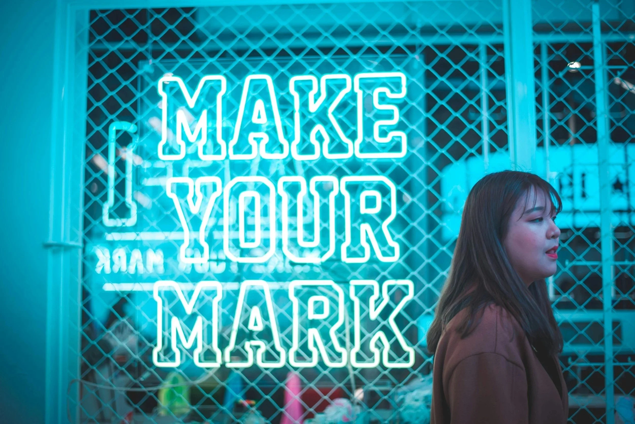 Neon sign reading 'Make Your Mark' behind a woman with brown hair and brown jacket, standing in front of a storefront window with a grid pattern