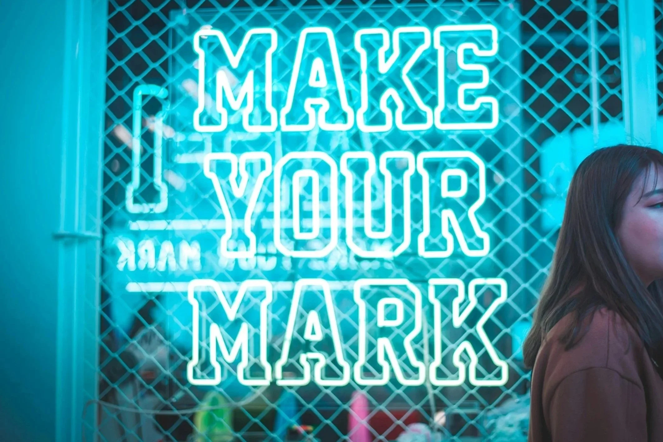 Neon sign with the message 'Make Your Mark' in bright blue letters on a wire grid background, with part of a woman's head and shoulder visible on the right side.