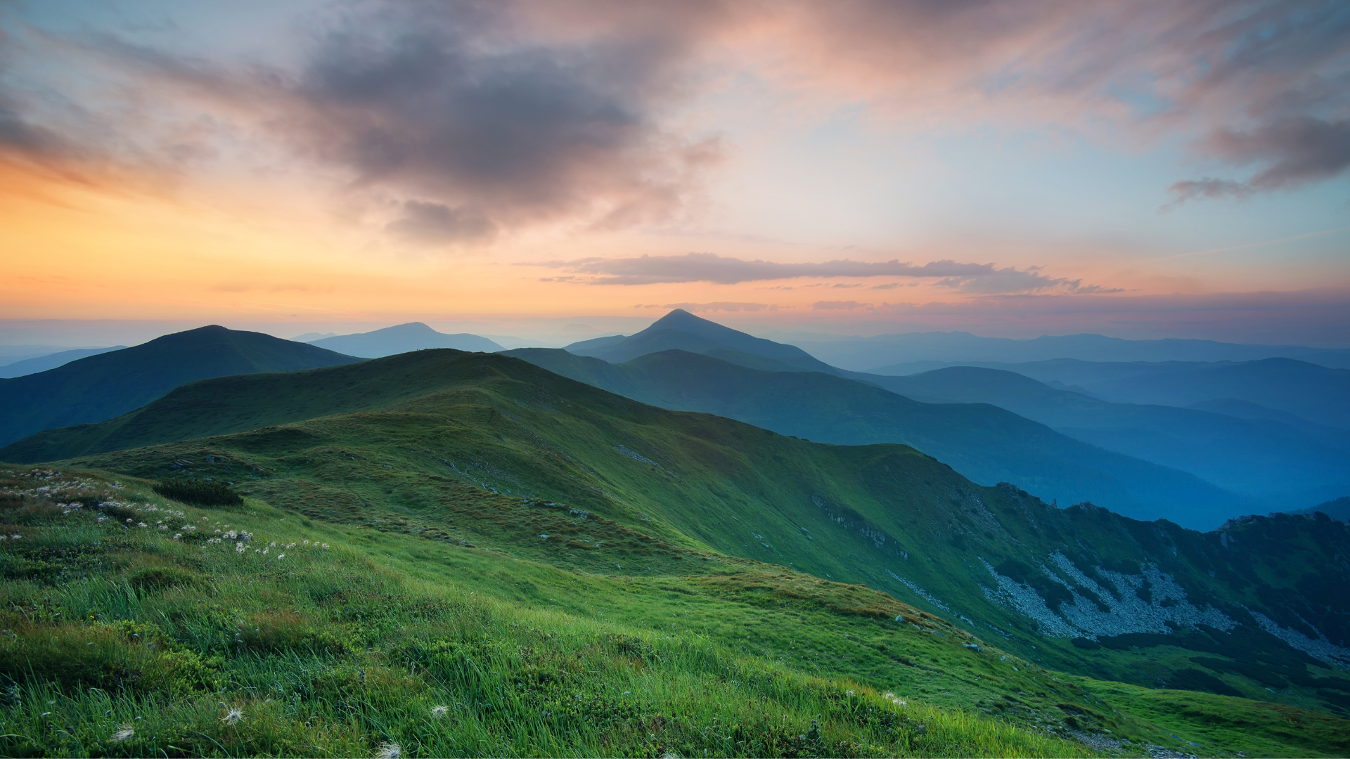 Sunset over green rolling mountains with layers of distant peaks and a partly cloudy sky.