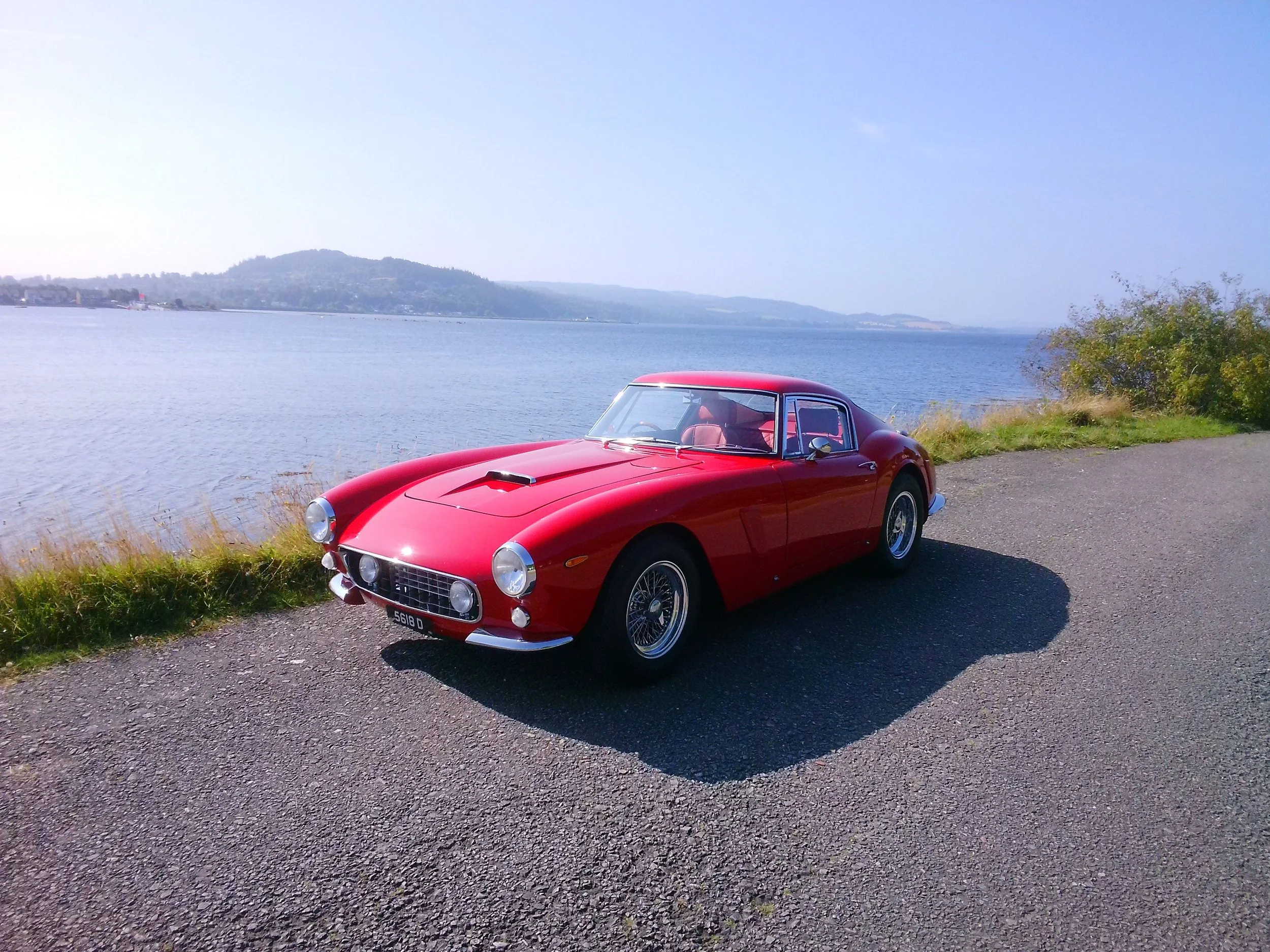 A vintage red sports car parked on a paved area near a large body of water with hills in the background and some greenery on the side.